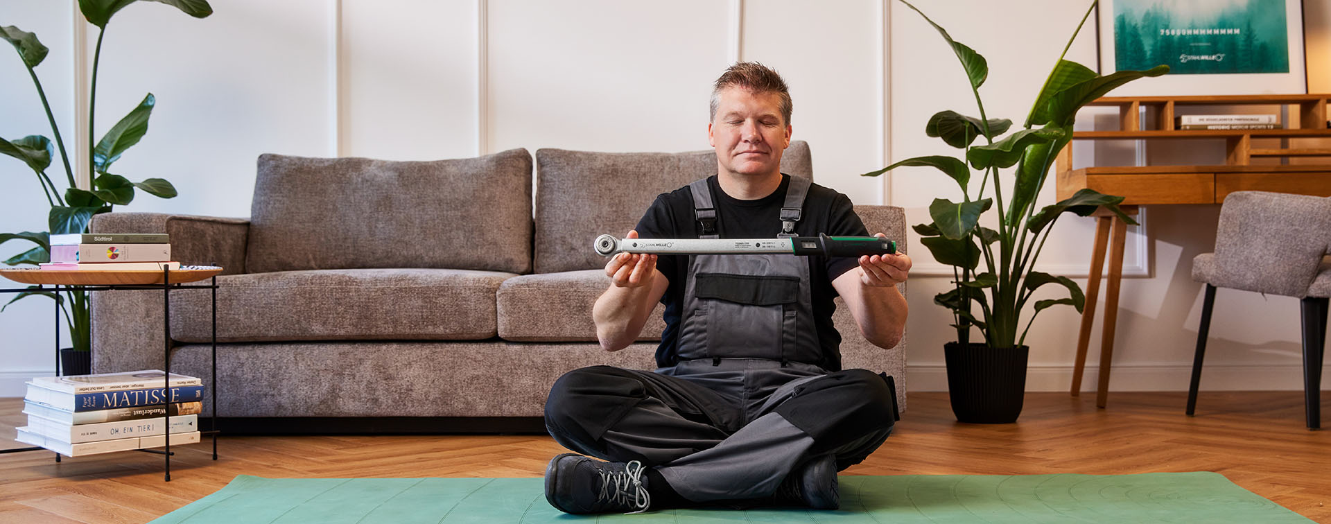 Mann sitzt in einem Wohnzimmer auf einer Yogamatte und hält einen Drehmomentschlüssel in der Hand