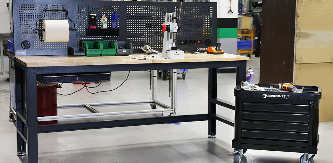 STAHLWILLE WB 630 werkbank in een professionele werkplaats – met massief houten werkblad, geïntegreerde stekkerblok met USB-aansluiting, opbergbakken en papierrolhouder. Ernaast staat een mobiele STAHLWILLE Top Box voor maximale flexibiliteit en organisatie.