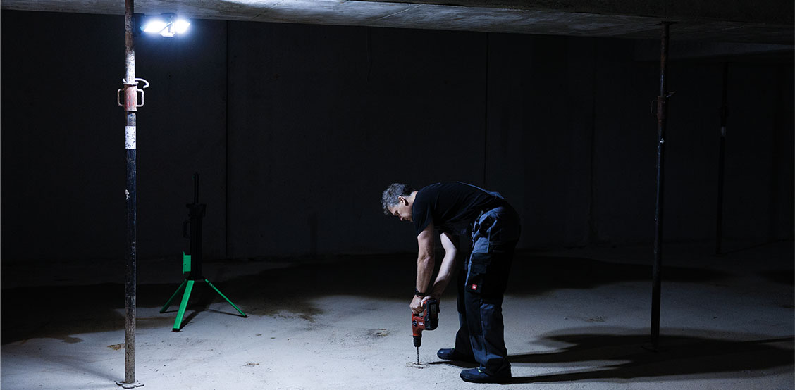 Ein Handwerker arbeitet mit einer Bohrmaschine in einem dunklen Kellerraum. Über ihm sorgt ein heller LED-Arbeitsstrahler auf grünem Stativ für großflächige Ausleuchtung.